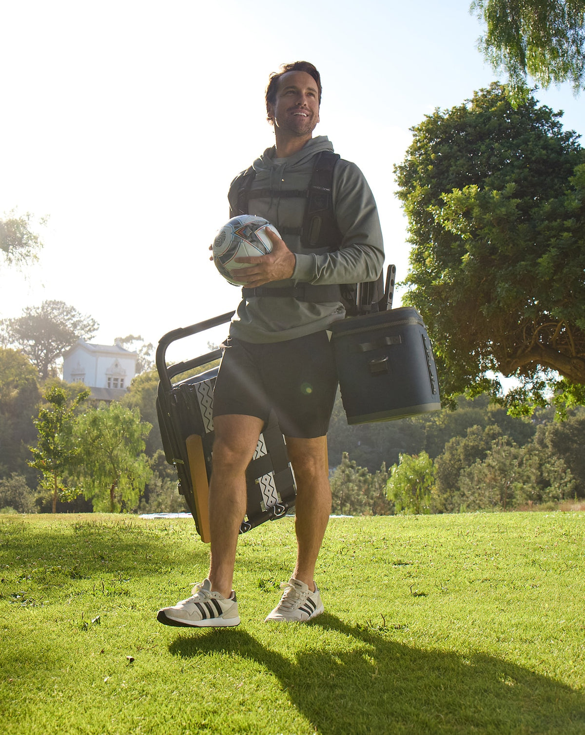 A man standing on a sunny grassy lawn outdoors, wearing a gray hoodie, black shorts, and white sneakers, while wearing the Hands Down Backpack - keeping him hands free as the backpack is carrying his cooler and lawn chair.